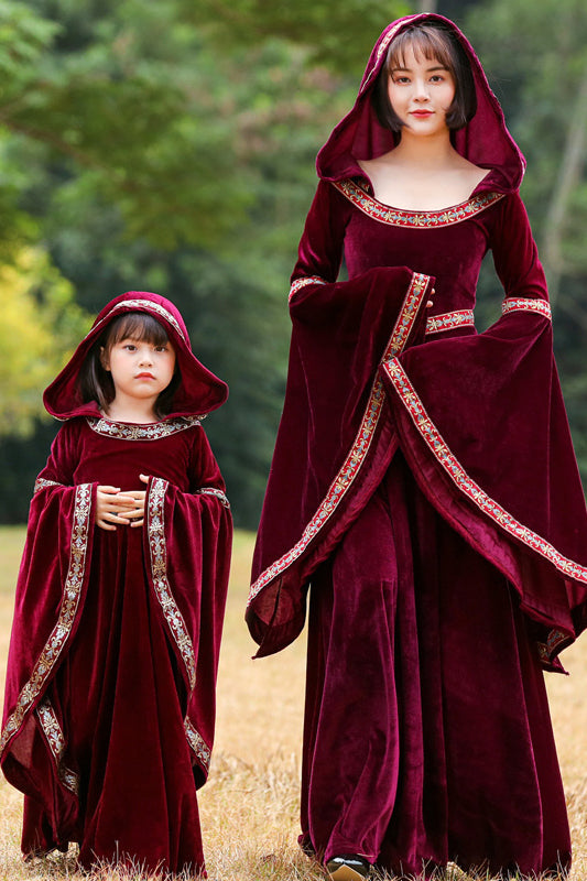 A woman and a young girl wearing matching medieval-style burgundy velvet hooded robes with ornate gold and silver embroidered trim, standing outdoors in a grassy field with a blurred green forest background.