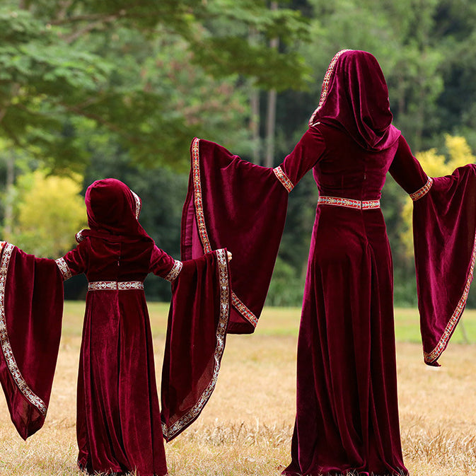 mom and daughter in matching deep burgundy velvet hooded maxi gowns with wide draped sleeves and ornate gold-trimmed borders, standing in a field with trees in the background.