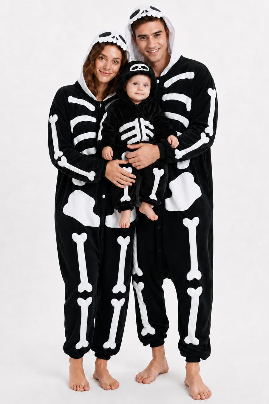 A family of three wearing matching black skeleton-themed hooded onesies, standing barefoot against a plain white studio background.